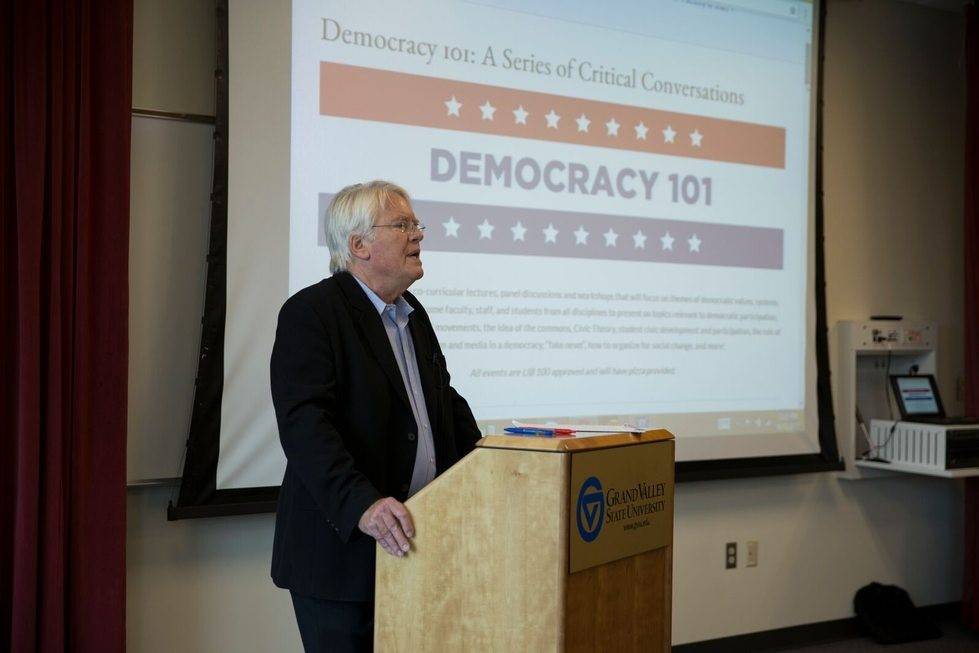 A speaker presenting to a room with a screen behind him that reads "Democracy 101"